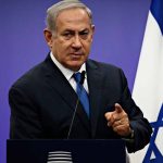 Man in suit giving speech in front of Israeli flag.