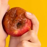 Person biting a rotten apple with yellow background