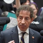 Man in suit surrounded by microphones and protesters