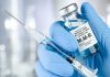 a hand in a blue nitrile glove holds a syrgine and a bottle of MMR vaccine