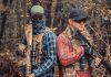 Two armed individuals in tactical gear in forest.