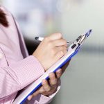 a supervisor in a pink jacket makes marks on a clipboard with a pen.