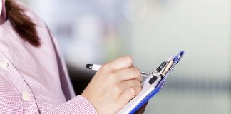 a supervisor in a pink jacket makes marks on a clipboard with a pen.