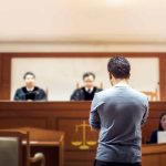 Man stands in courtroom before judges and stenographer
