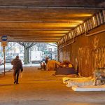 People walk under a bridge with homeless encampment