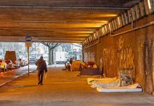 People walk under a bridge with homeless encampment