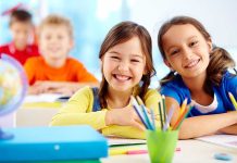 Two smiling girls in a classroom with colorful stationery and classmates in the background