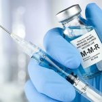 Measles Discovery Raises Concerns Over Global Travel Health Risks a hand in a blue nitrile glove holds a syrgine and a bottle of MMR vaccine