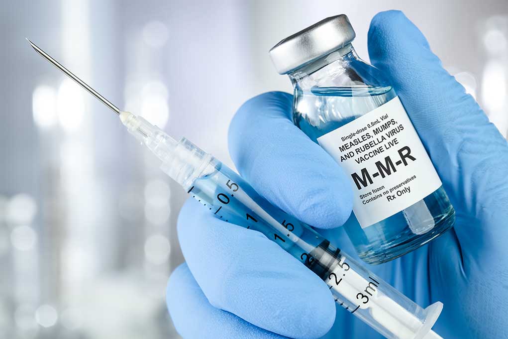 1449408860 featured image a hand in a blue nitrile glove holds a syrgine and a bottle of MMR vaccine