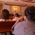 High-Profile Pastor’s Pending Arrival Stirs Controversy In Oklahoma People sitting in church pews during service.