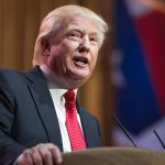 Details Emerge From July Trump Assassination Attempt Sparking Further Questions Man in suit speaking at podium with flag background