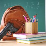 Arkansas Schools Introduce Controversial New Course To Boost Student Safety Gun and backpack on desk with school supplies.