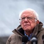 Bernie Sanders Signals Support For Joe Rogan’s Health Reform Dialogue Elderly man in jacket speaking at outdoor event.