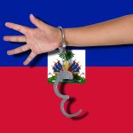 Haiti Unveils “War Budget” To Tackle Gangs person's arm with handcuffs in front of the flag of Haiti