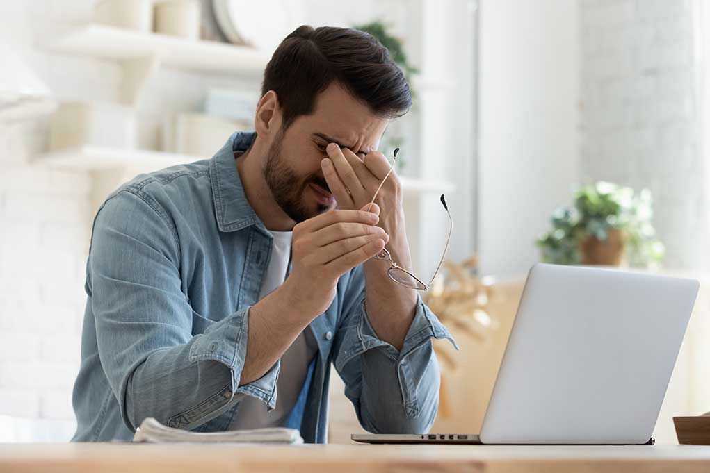 Man rubbing eyes holding glasses near laptop