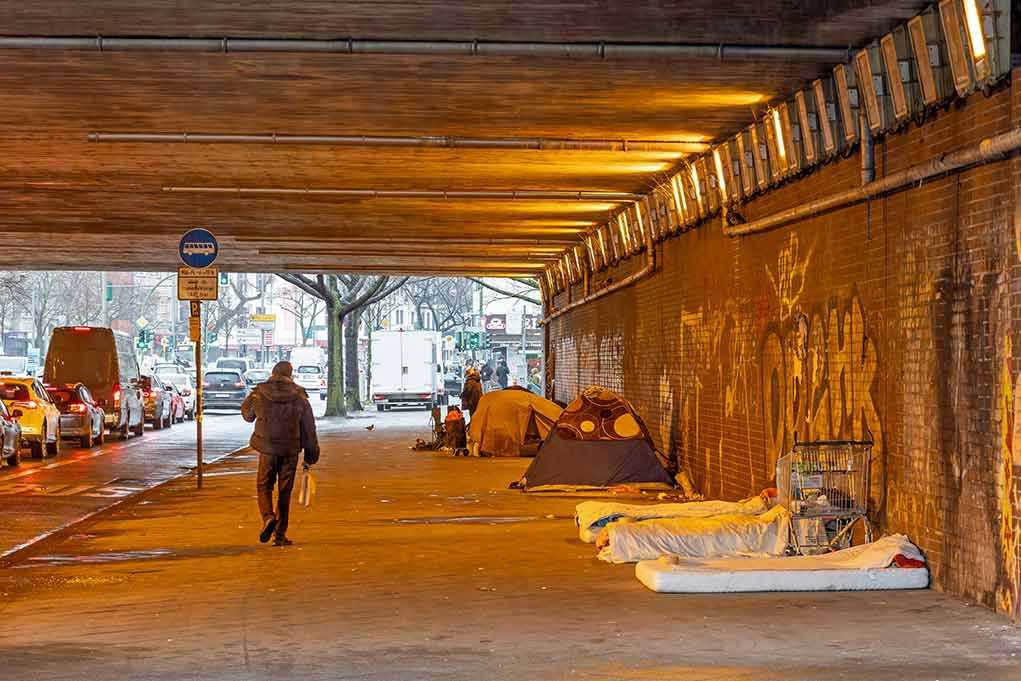 2423598655 People walk under a bridge with homeless encampment