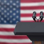 Podium with microphones in front of American flag.
