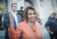 Woman in orange blazer speaking, surrounded by security personnel.