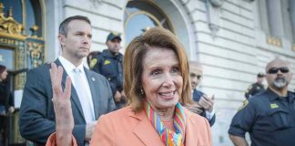 Woman in orange blazer speaking, surrounded by security personnel.
