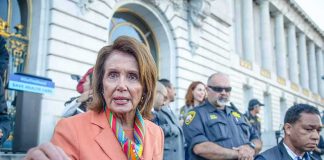 A woman in an orange blazer speaking passionately at a rally with security personnel in the background