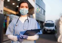 Nurse in scrubs and mask outside hospital holding clipboard.