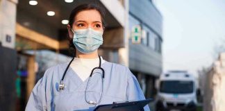 Nurse in scrubs and mask outside hospital holding clipboard.