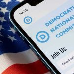 Massive DNC Money Laundering Fraud EXPLODES – See Report! Phone displaying Democratic National Committee website over flag.
