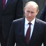Putin’s Audacious U.S. Expert Buyout Attempt A serious-looking man in a suit walking outdoors with security personnel in the background