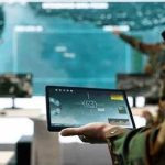 Soldier holding a tablet in a command center with digital maps on screens
