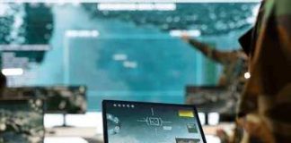 Soldier holding a tablet in a command center with digital maps on screens
