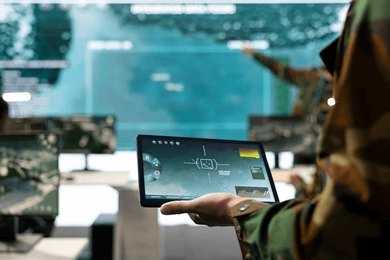 Soldier holding a tablet in a command center with digital maps on screens