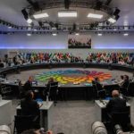 A large round table meeting with global leaders and flags in the background
