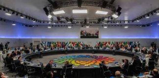 A large round table meeting with global leaders and flags in the background