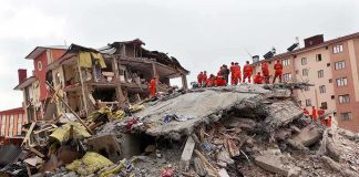 Drone Barrage Torches Homes — Elites Yawn Rescue workers in orange uniforms on a collapsed building site after an earthquake