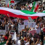 Iran’s Supreme Leader Dies—Now What? Crowd of protesters holding signs and a large tricolor flag
