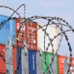 Midnight Tariff Ambush Shocks 60 Nations Razor wire fence in front of stacked shipping containers