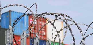 Midnight Tariff Ambush Shocks 60 Nations Razor wire fence in front of stacked shipping containers