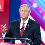 Spy-Tainted Money Haunts California Power Man in suit speaking at a podium on a colorful stage