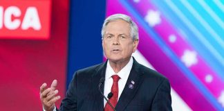 Spy-Tainted Money Haunts California Power Man in suit speaking at a podium on a colorful stage