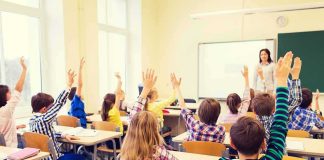 Students raise hands in a classroom with a teacher at the front